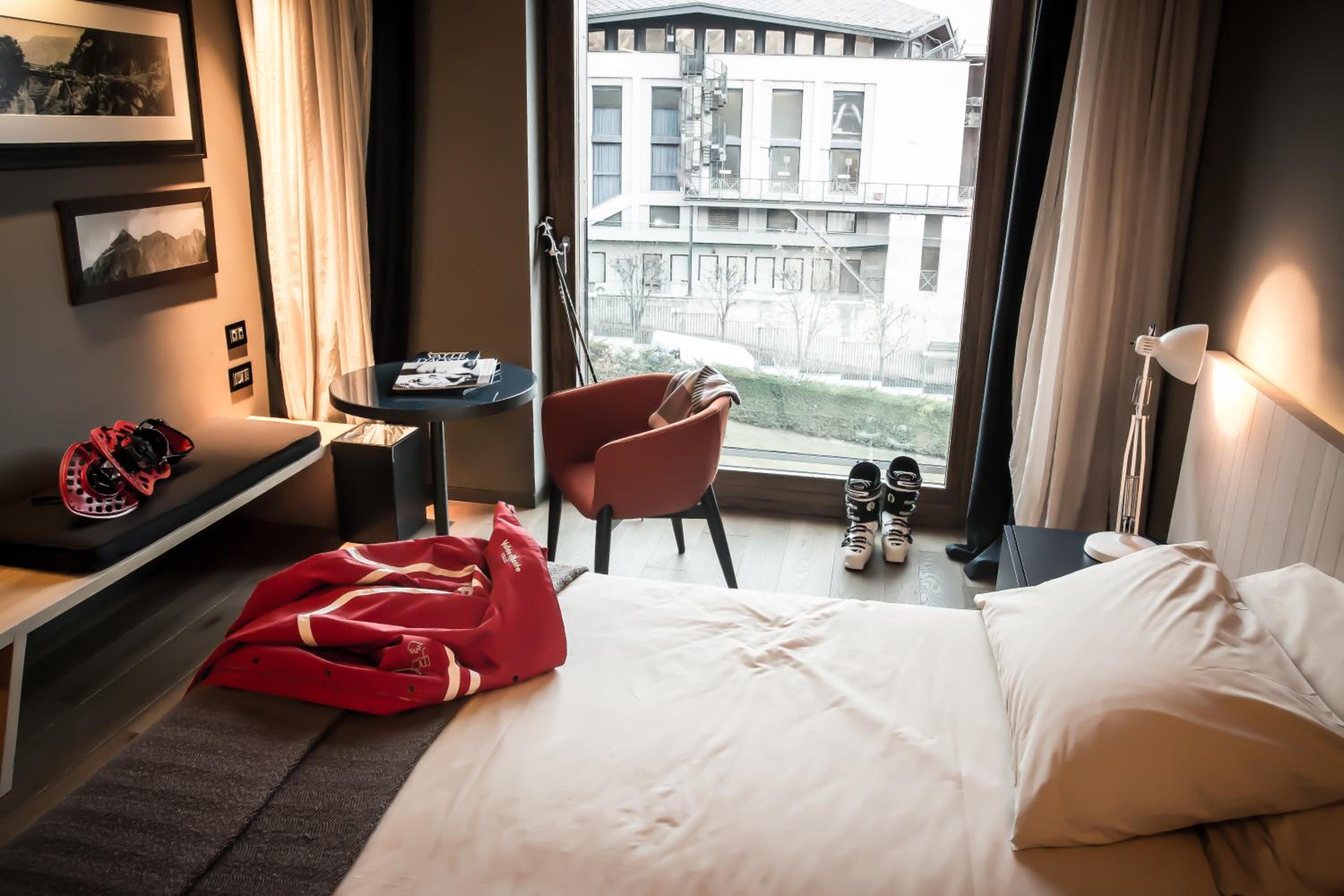 Hotel room with a white bed, red chair, and large window overlooking a building.