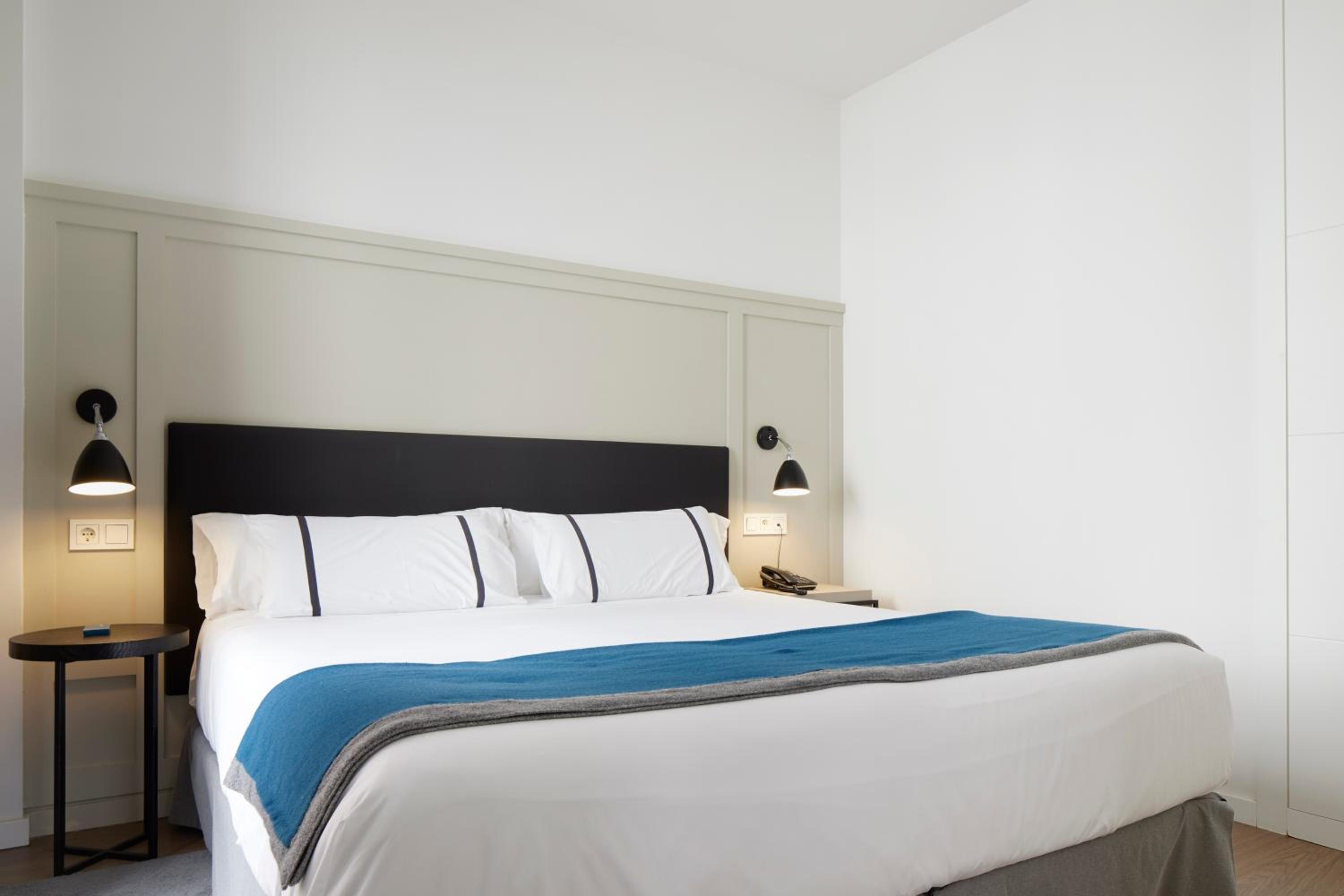 Modern hotel bedroom with large white bed, blue throw, black headboard and wall lamps