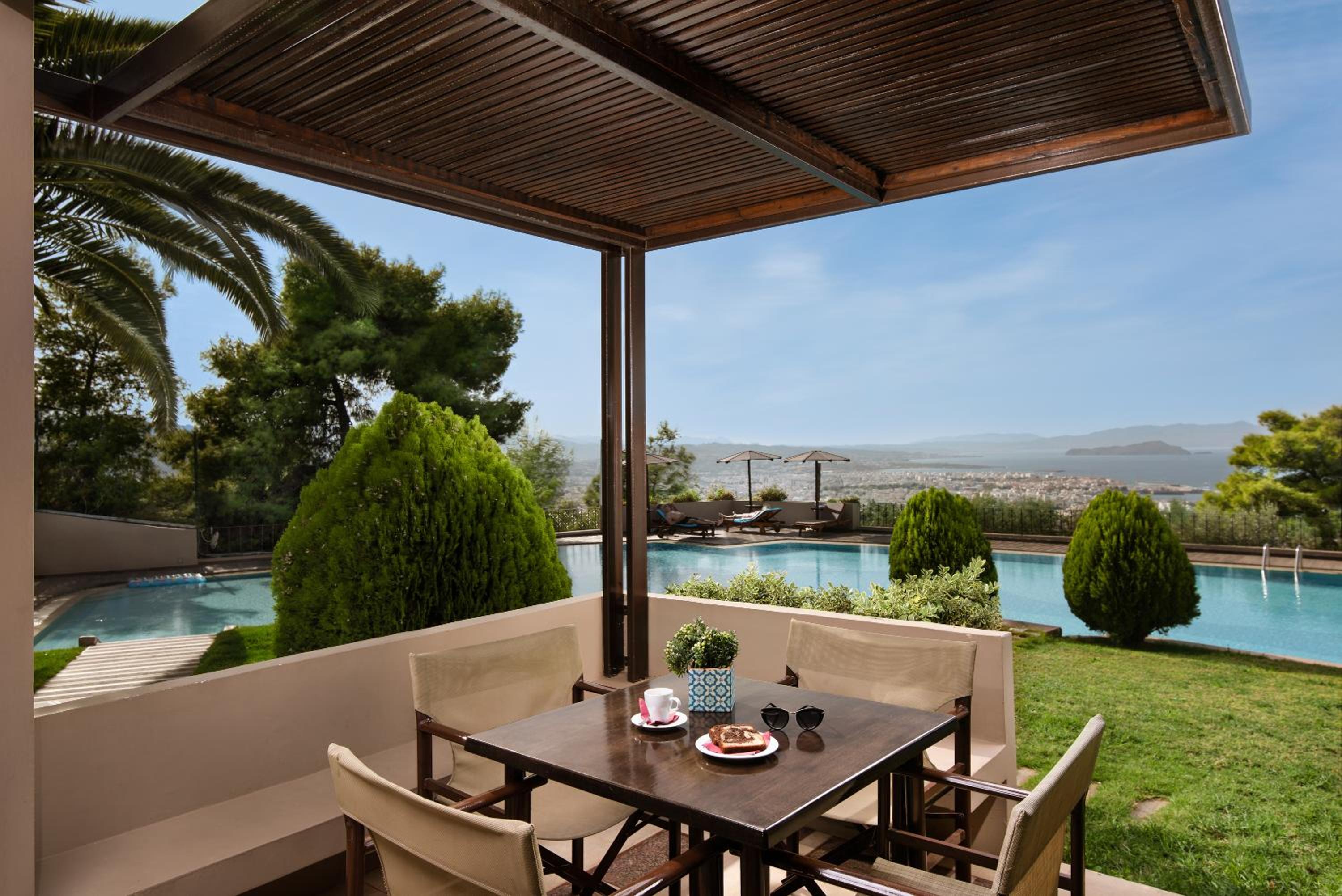 Covered balcony with a dining table and chairs overlooking a swimming pool and distant sea view.