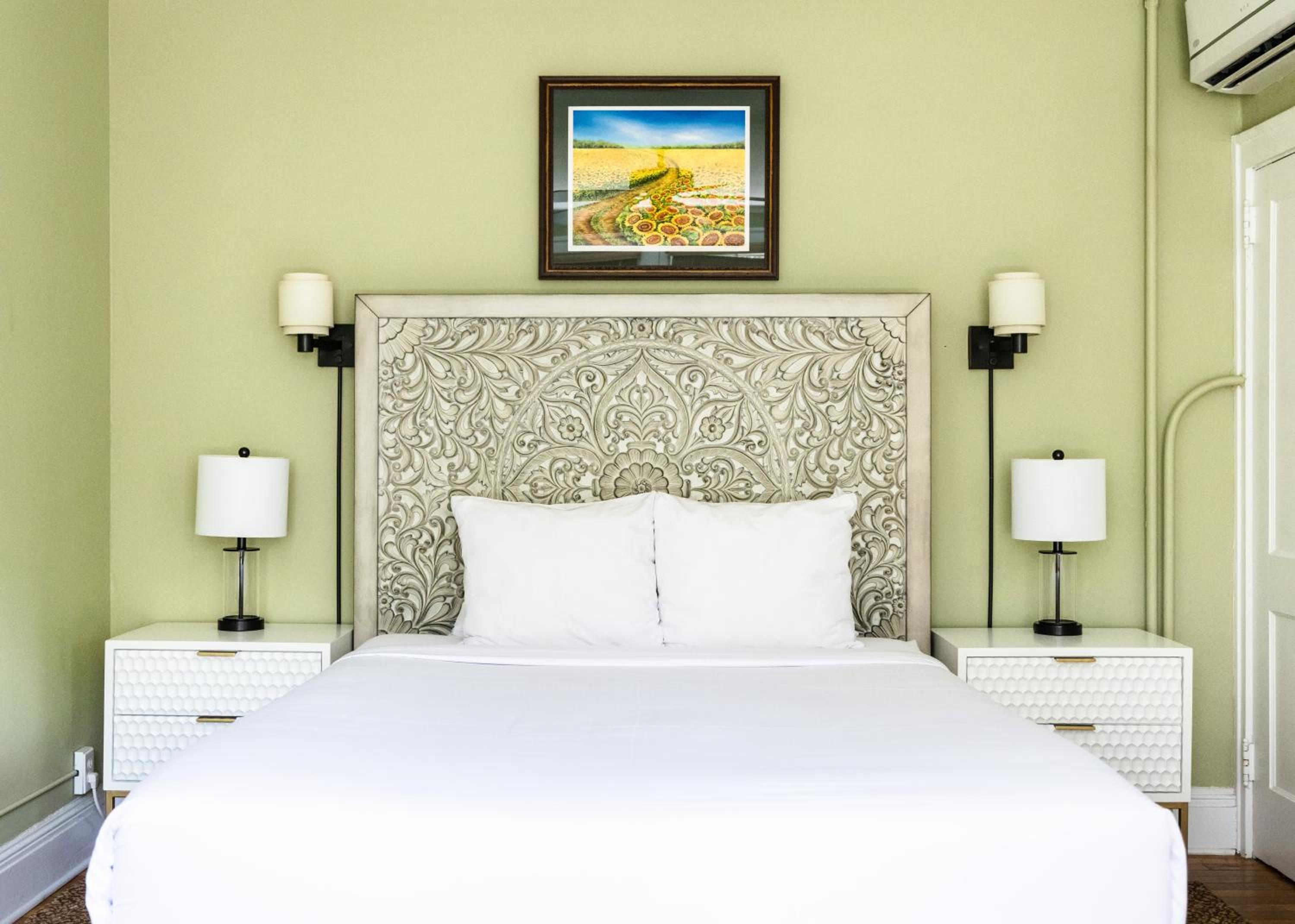 Bed with carved headboard, white linens, nightstands and sunflower painting on green wall