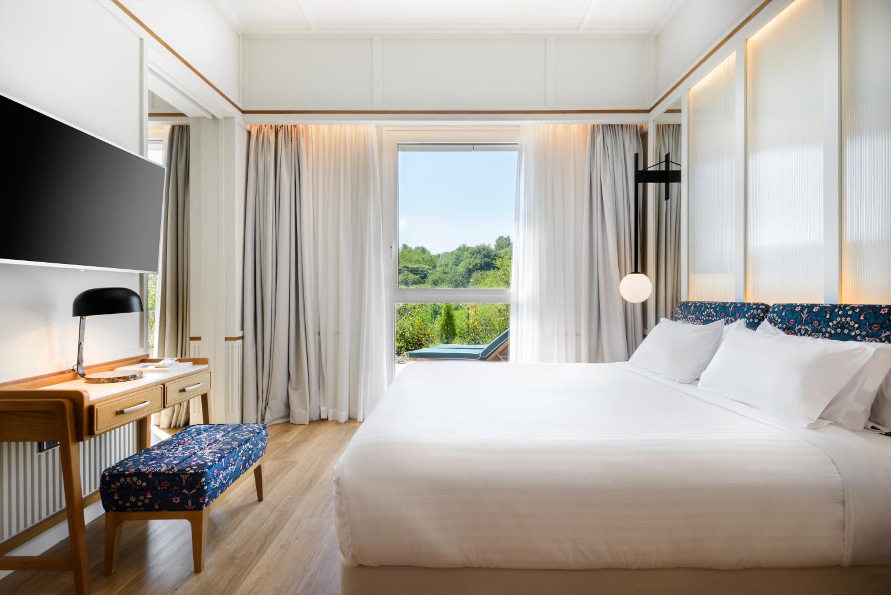 Hotel room with a large bed, wooden desk, and a window overlooking a garden.