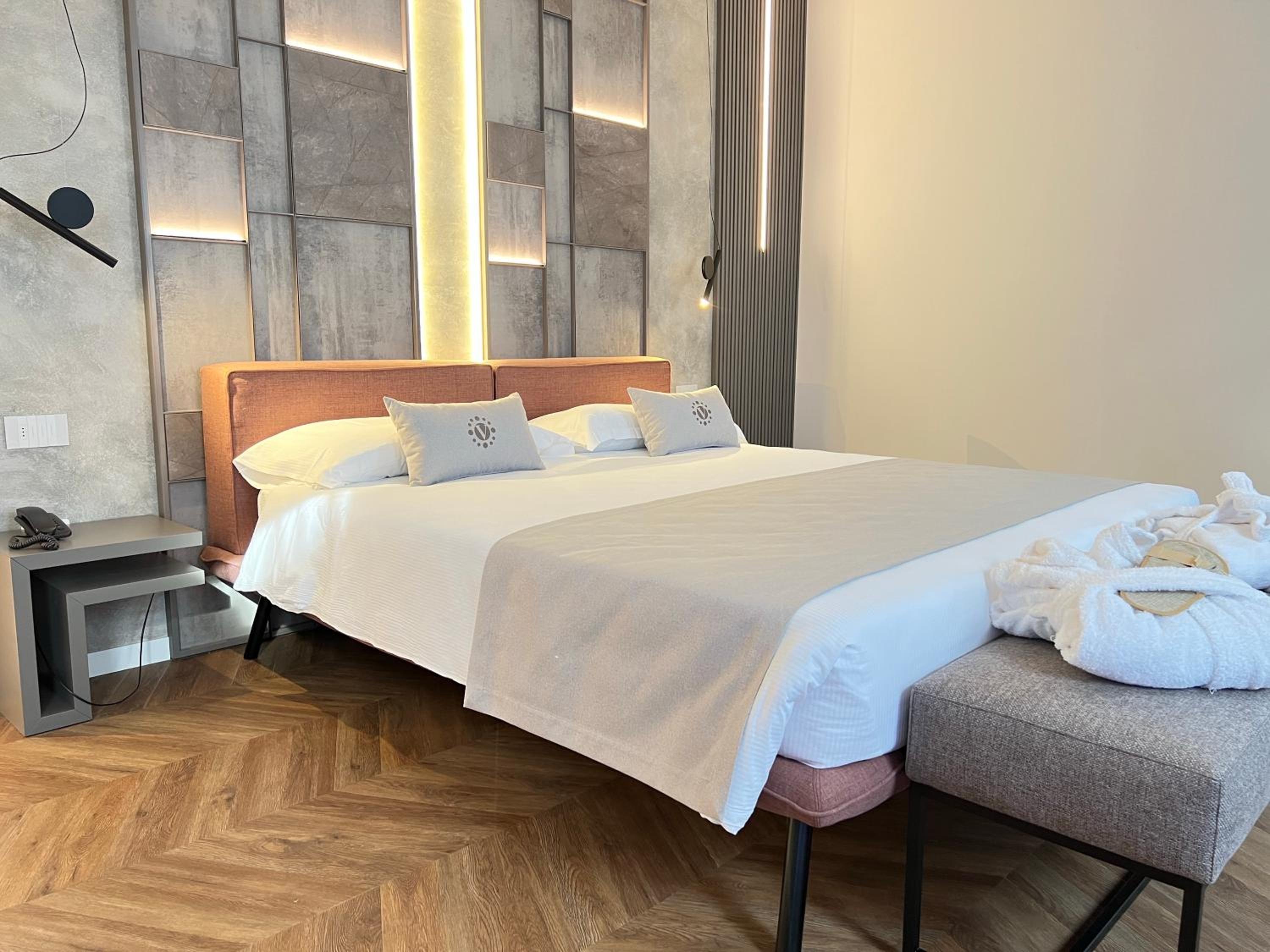 Modern hotel bedroom with large bed, lit stone-look headboard wall, bathrobes on bench