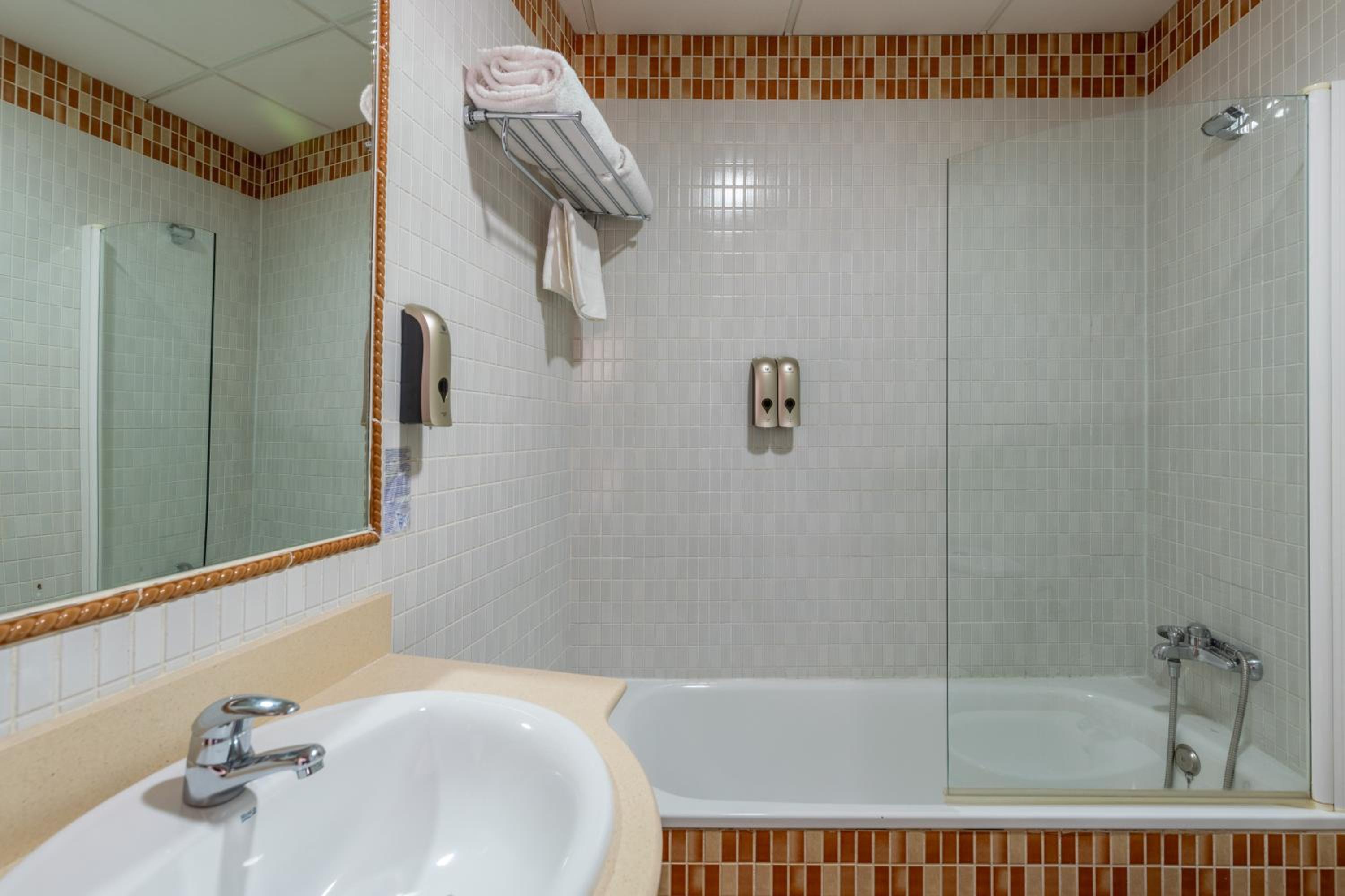 Tiled hotel bathroom with sink, bathtub with glass screen, towel rack and soap dispensers