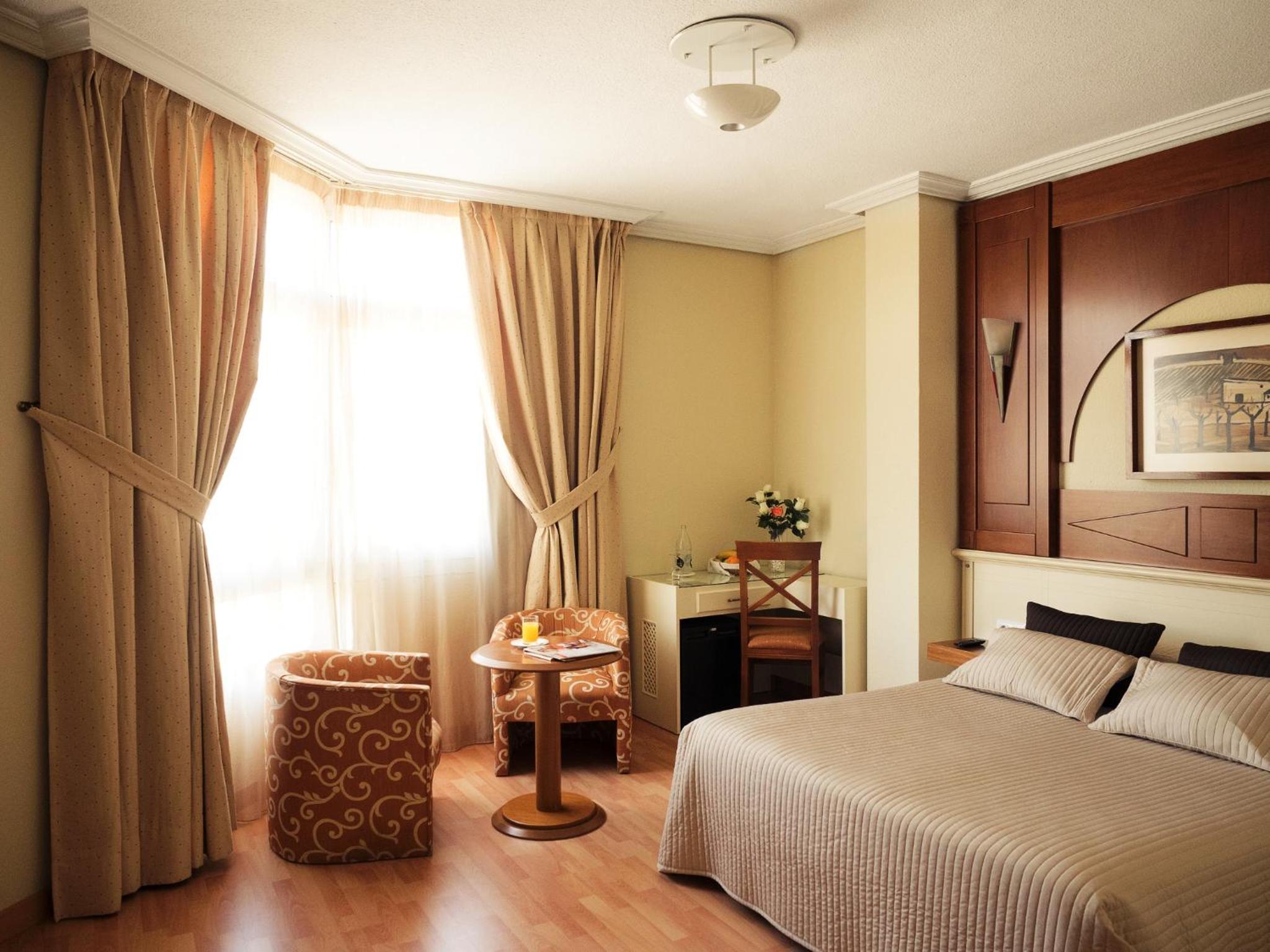 Hotel room with a double bed, patterned armchairs, and a large window with beige curtains.