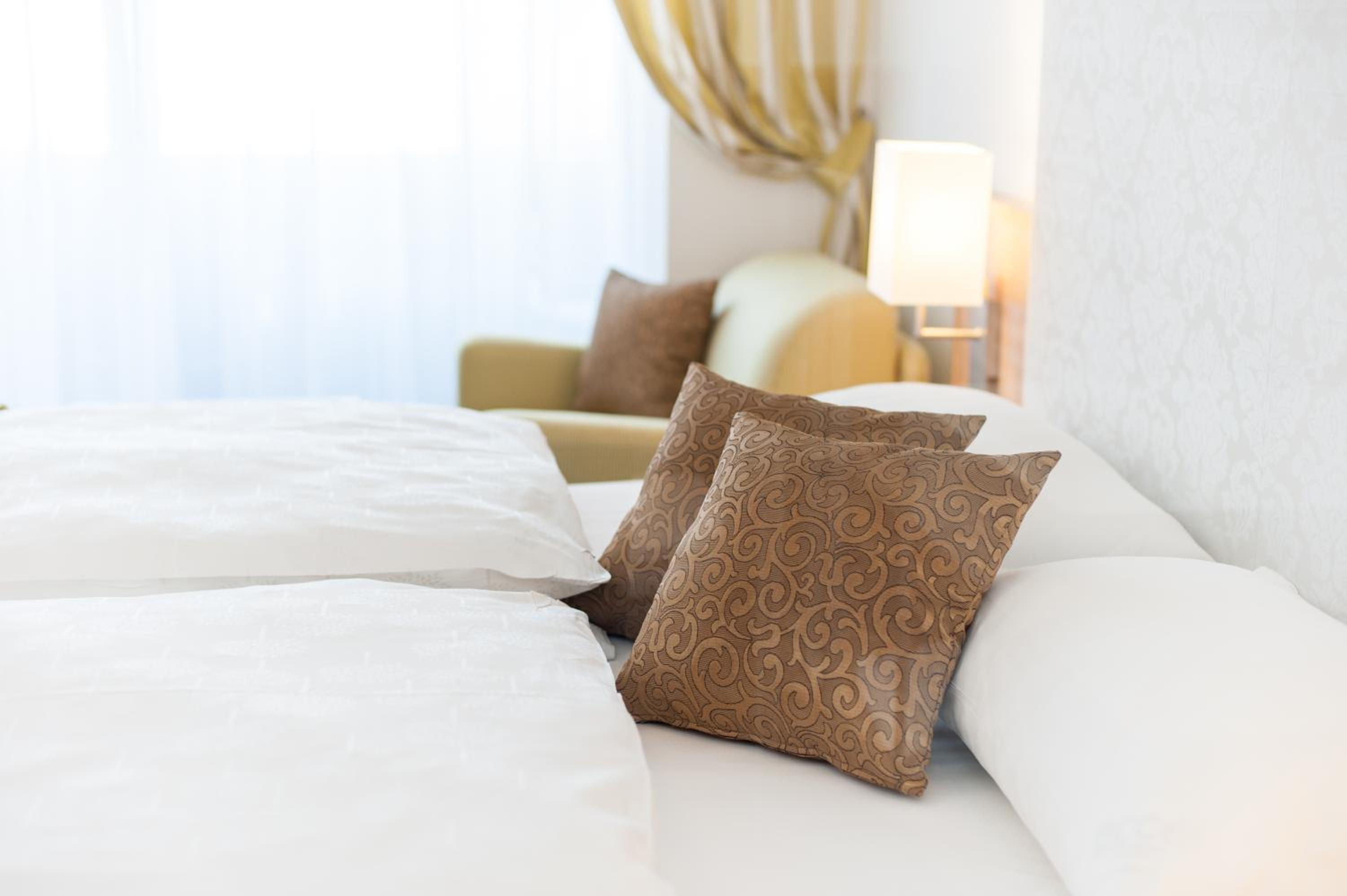 White bed with brown patterned cushions, lamp, curtain and armchair behind