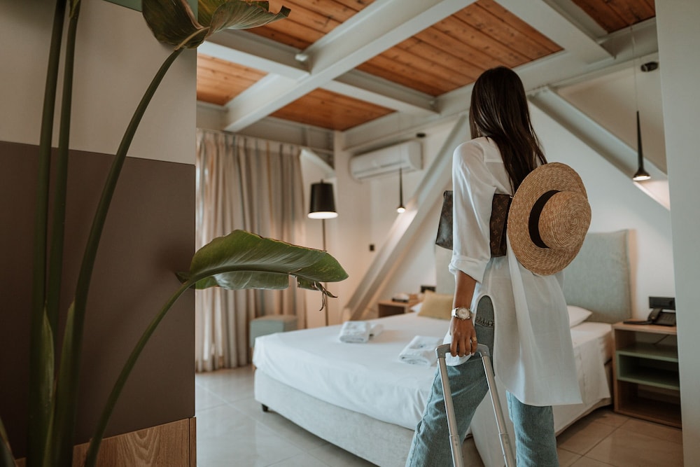 Hotel bedroom with a white bed, wooden ceiling beams, and a large potted plant.